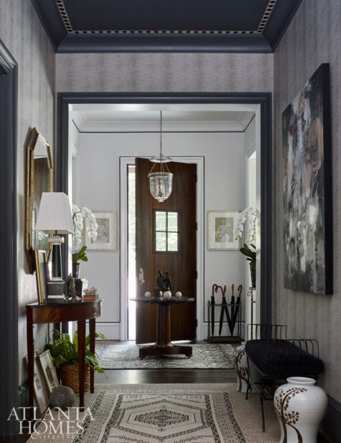 Entryway with gray patterned wallpaper, dark wood door, ornate rug, and console table with lamp and plants.