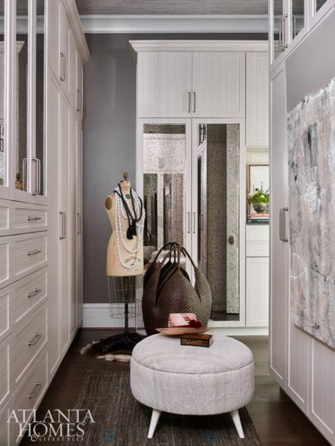 Walk-in closet with floor-to-ceiling white cabinetry, mirrored doors, a mannequin with necklaces, a large woven tote bag, and a round ottoman with white legs.