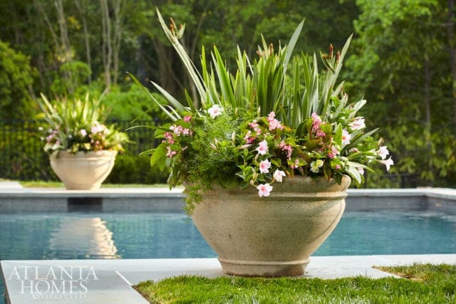 Large stone planter filled with pink flowers and tall green foliage sits beside a swimming pool with lush trees in the background.