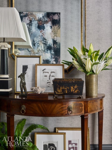 Mahogany console table with lion ring pulls, topped with a lamp, abstract art, framed sketches, a decorative box, and white lilies in a gold vase.