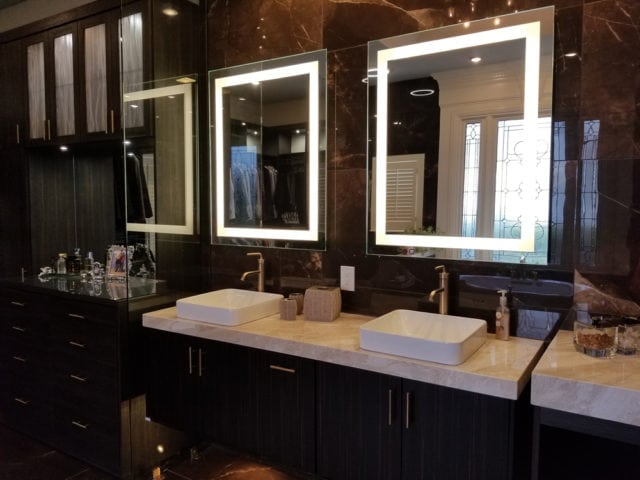 Dark wood vanity with marble countertop and two white vessel sinks, each with a backlit mirror and brass faucet. Dark marble walls and built-in cabinetry.