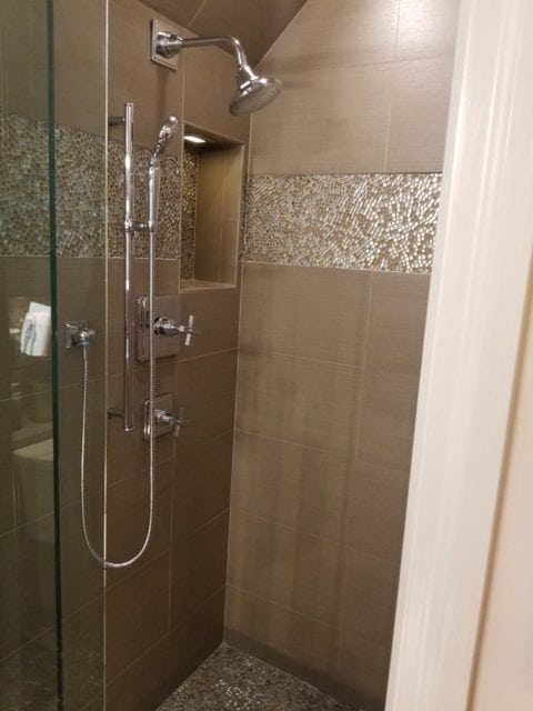 Shower stall with large format taupe tiles, a band of iridescent mosaic tile, a recessed niche, and a pebble tile floor. Chrome fixtures include a rain shower head and handheld wand.