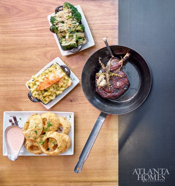 A steak in a pan, mac and cheese with lobster, broccoli with breadcrumbs, and onion rings with dipping sauce on a wooden table.