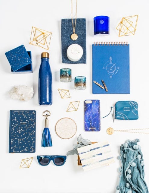 Flat lay of blue and gold constellation-themed items including a notebook, phone case, water bottle, jewelry, candles, sunglasses, and a tassel keychain.