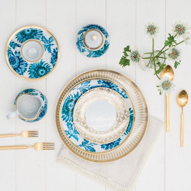 Gold-rimmed dinnerware set with blue floral and geometric patterns, including plates, a creamer, a sugar bowl, and gold cutlery, arranged on a white wooden surface.