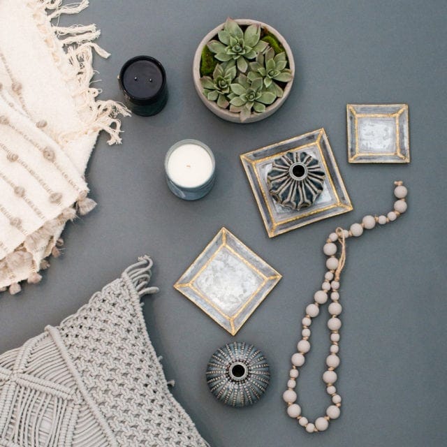 Flat lay of decorative items including a macrame pillow, textured throw blanket, two candles, a potted succulent, decorative frames, a beaded necklace, and two ornamental objects.