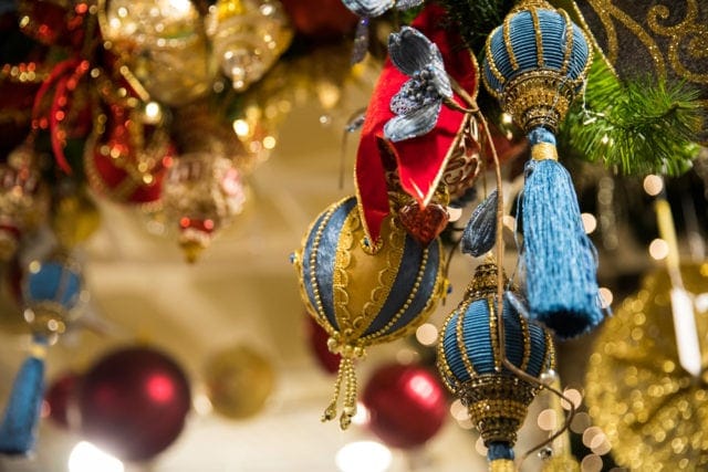 Close-up of ornate blue and gold Christmas ornaments with tassels hanging from a decorated tree with red ornaments and bokeh lights in the background.