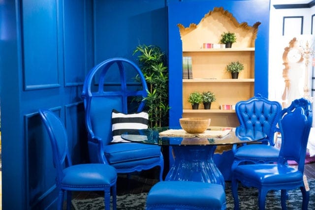 Blue dining room with ornate chairs, a glass-top table, and a wooden shelf unit displaying plants and books.