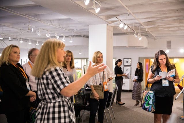 Group of people attending an event, with one woman in the foreground speaking and gesturing. Other attendees stand and listen, some holding papers.