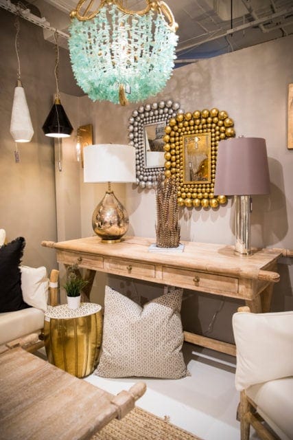 Light wood console table with three drawers, topped with two lamps, a vase of feathers, and two decorative mirrors. A large aqua chandelier hangs above.