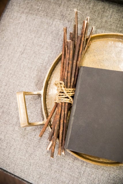 Bundle of thin, dry twigs tied with rough twine sits on a brass tray next to a closed black book on a textured gray surface.