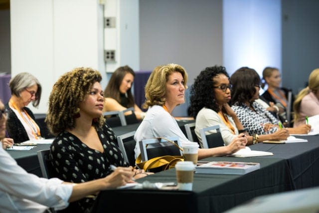 A diverse group of women attend a conference, seated at tables and listening attentively.