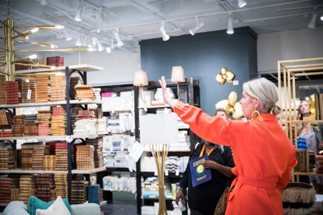 Woman in orange jacket gesturing towards shelves filled with books and decorative items in a retail space.