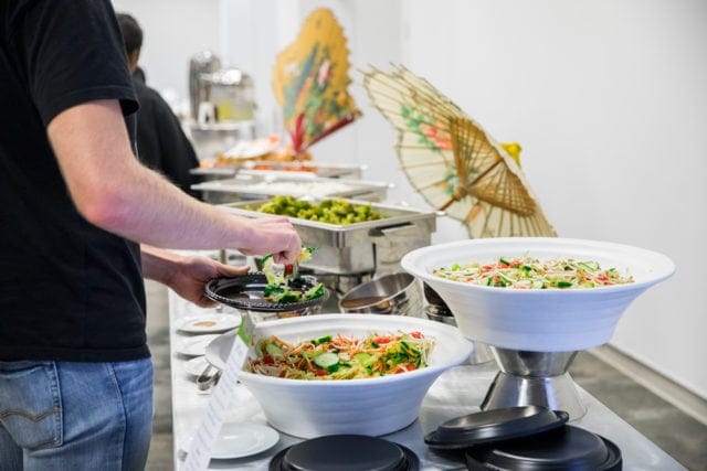 Person serving salad from a buffet line with chafing dishes and decorative parasols in the background.
