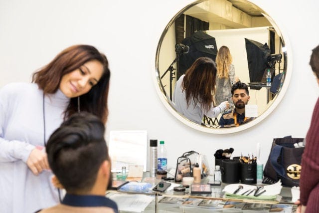 A woman with dark hair applies makeup to a man with dark hair in a studio setting, reflected in a large round mirror.