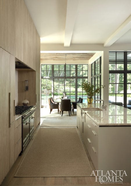 Forming the visually pleasing planes of the kitchen are custom white oak Karpaty Cabinets featuring the same undulating texture as the fireplace on the opposite side of the room. Here, a Waterworks tile backsplash repeats this linear motif for a seamless effect. The sconce by the stove is from The Urban Electric Co.