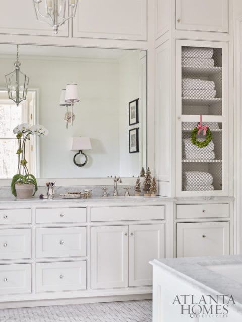 White bathroom vanity with marble countertop, silver hardware, and a large mirror reflecting wall sconces and a chandelier. A glass-front cabinet displays folded white towels and a small wreath.