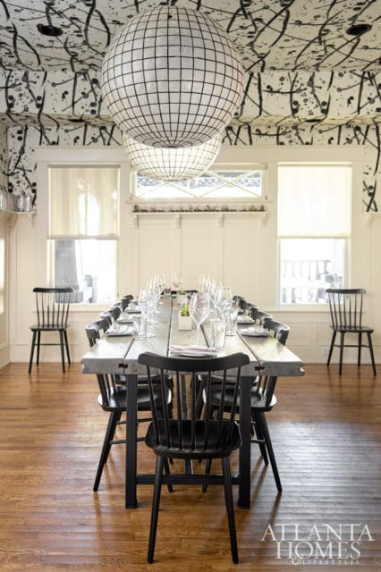 Dining room with a long wooden table set for a meal, black spindle-back chairs, and two large spherical pendant lights hanging from a ceiling with black splatter-pattern wallpaper.