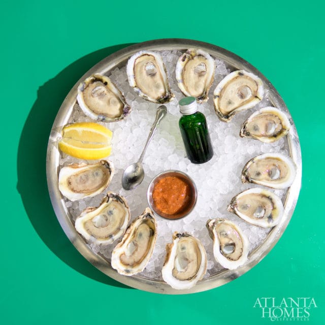 A silver platter filled with ice and a dozen oysters, served with lemon wedges, a small dish of cocktail sauce, a spoon, and a small green bottle.