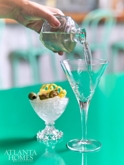 A hand pours clear liquid from a glass decanter into a martini glass filled with ice, garnished with pineapple and an olive.