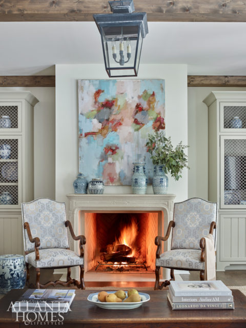 Built-in bookcases with lattice detailing flank the limestone fireplace by Francois & Co., which is topped by a lively abstract painting. “We wanted the family den to blend seamlessly with the look of the kitchen and breakfast room, so we used the same fabrics in soft neutral shades of blue and taupe,” says DeLoach.