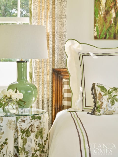 Bedroom with white bedding, green and brown striped trim, floral accent pillow, green lamp on glass table, and patterned curtains.