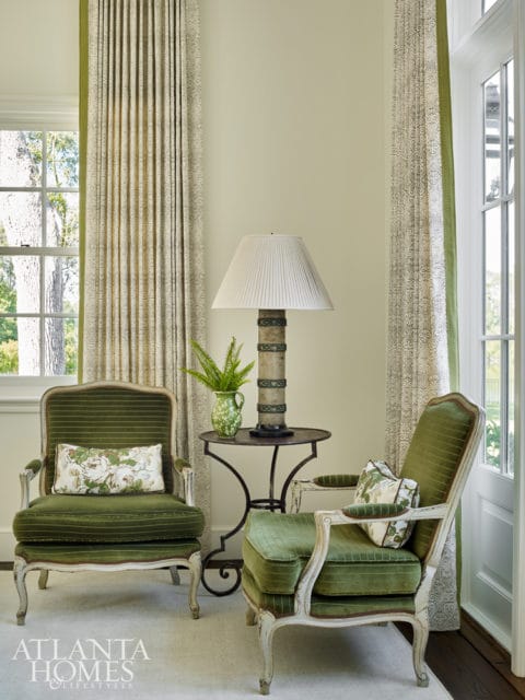 Two antique green velvet armchairs flank a small metal side table with a lamp and vase. Patterned curtains hang on either side of the window.