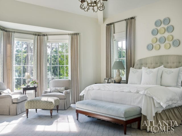 Bedroom with a king-size bed, tufted headboard, and white linens. A light blue bench sits at the foot of the bed. Two upholstered armchairs and an ottoman are arranged near a bay window.
