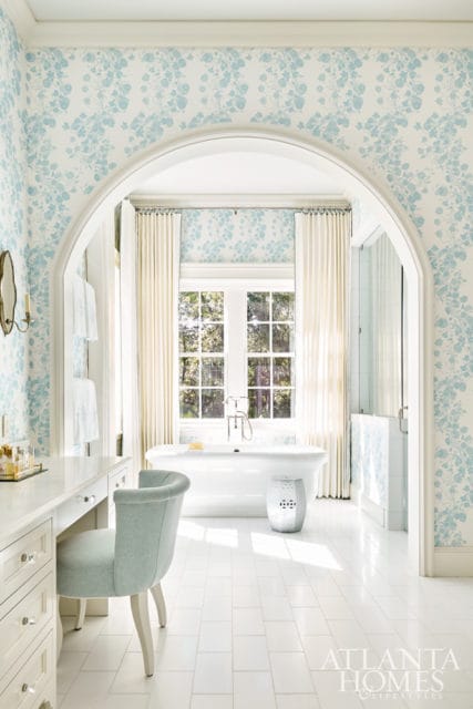 Light blue floral wallpaper adorns this bathroom featuring a freestanding white tub, a vanity with a light blue upholstered chair, and white tile flooring.