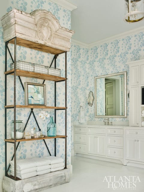 White bathroom with blue floral wallpaper, a tall open shelf unit with decorative wood top, and a white vanity with a silver mirror