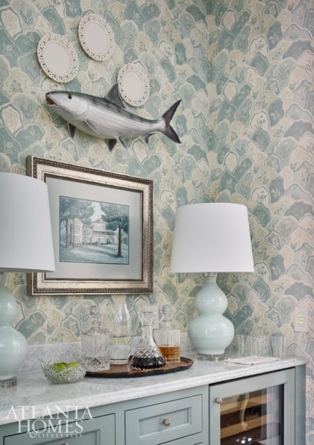 Bar area with blue cabinetry, marble countertop, and light blue lamps. A taxidermied fish and framed artwork hang on a blue and cream patterned wallpaper.