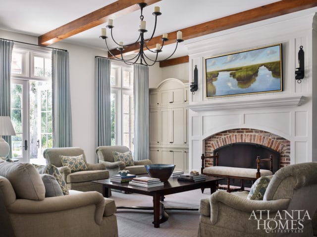 Living room with exposed wooden beams, a large brick fireplace with a painting above, and upholstered furniture.