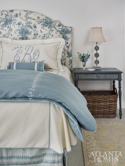 Bedroom with a blue floral upholstered headboard, white bedding with blue embroidered monograms and trim, a light blue duvet, and a dark blue bedside table with a lamp and basket.