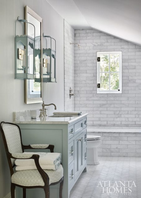 Light blue vanity with marble countertop, silver faucet, and white subway tile walls in a bathroom with a herringbone tile floor and a window overlooking trees.