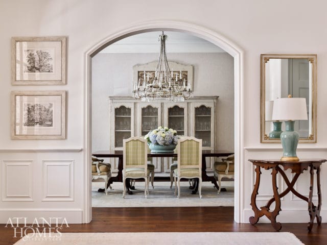 View through an arched doorway into a dining room with a crystal chandelier, antique china cabinet, and upholstered chairs. A console table with a lamp and mirror is visible in the foreground.