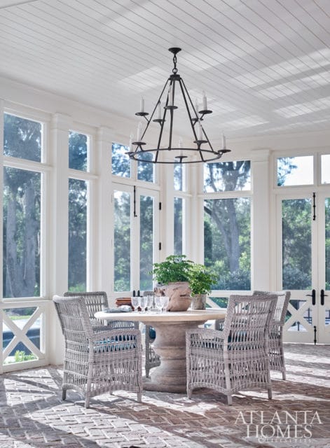 Sunroom with white paneled ceiling, large windows overlooking trees, and a round stone pedestal table with four gray wicker chairs.