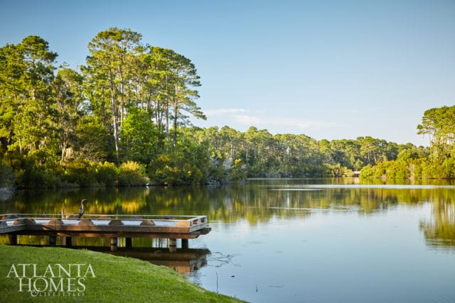 Wooden dock on a calm lake with a bird standing on it, surrounded by lush green trees and a clear blue sky.