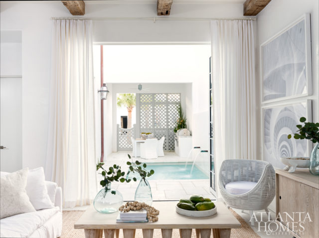 A pair of retractable windows—concealed by Ulster Linen drapery fabric—connect the breezy living room to the courtyard pool. A Cisco Brothers sofa and rattan chair from Bali offer chic spots for socializing or enjoying the views. Stone-like concrete tile flooring by Peacock Pavers feature porous properties and are designed to help catch sand.