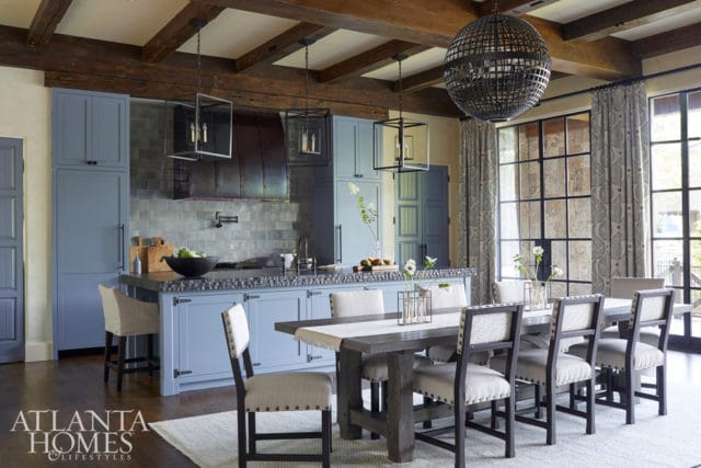 Blue kitchen island and cabinetry with a dark stone countertop, gray subway tile backsplash, and a rustic wooden dining table with upholstered chairs.