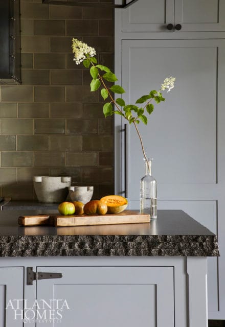 Gray kitchen with dark subway tile backsplash, stone countertop, and light gray cabinets. A cutting board with tomatoes and melon sits on the counter next to a glass vase with white flowers.