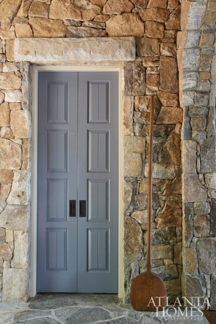 Double gray doors with paneled detailing set within a rustic stone wall, featuring a large wooden paddle leaning against the wall and a stone floor.