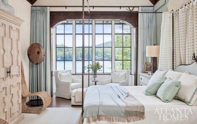 Bedroom with large windows overlooking a lake, featuring white upholstered chairs, a four-poster bed with light blue and beige throws, a light wood armoire, and a woven chair.
