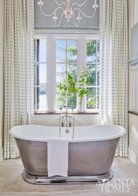 Freestanding bathtub with silver exterior and white interior, set against a large window with lake and tree views. White patterned curtains frame the window, and a white chandelier hangs from the blue ceiling.