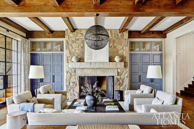 Living room with stone fireplace, exposed wooden beams, blue paneled doors, and neutral-toned furniture. A large orb chandelier hangs above the fireplace.