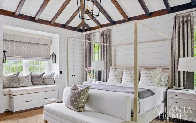 Bedroom with white shiplap walls, exposed wooden beams on the ceiling, a four-poster bed with patterned pillows, and a window seat with built-in storage.