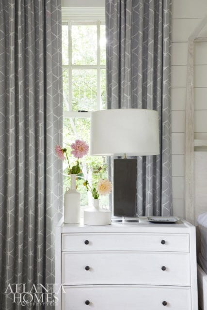 White dresser with black knobs next to a window with grey patterned curtains. A white lamp sits on the dresser with two vases of flowers.