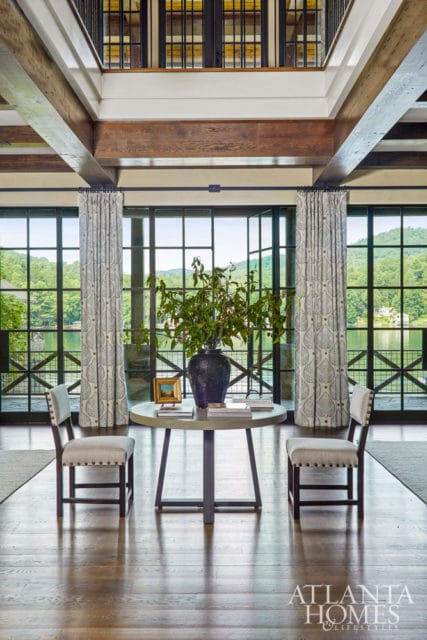 Foyer with exposed wooden beams, large windows overlooking a lake, a round table with a vase of greenery, and two upholstered chairs.