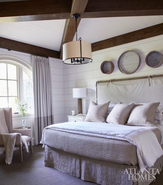 Bedroom with shiplap walls, exposed wood beams, a large bed with textured linens, and a window with arched top and draped curtains.