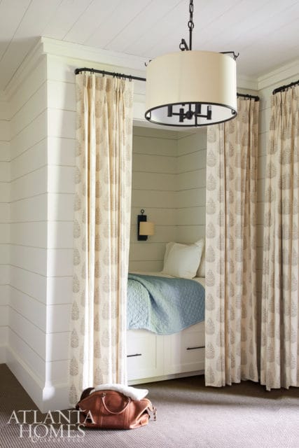 Built-in bed nook with shiplap walls, patterned curtains, blue quilted bedspread, and a drum pendant light overhead. A leather duffel bag rests on the carpet in the foreground.