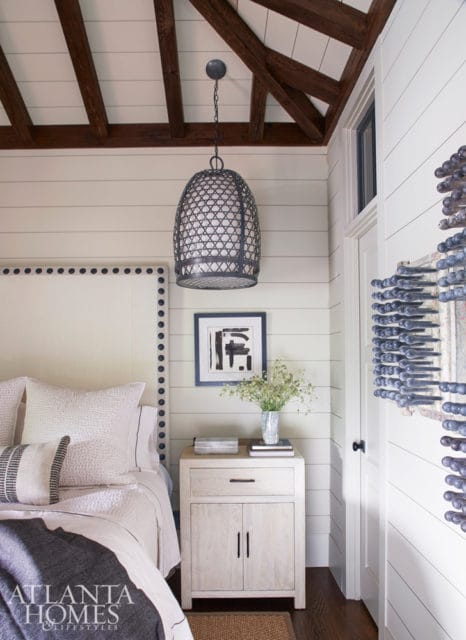 Bedroom with shiplap walls, exposed wooden beams on the ceiling, and a large pendant light. A white upholstered headboard with nailhead trim is visible, along with a nightstand holding a vase of flowers and artwork.